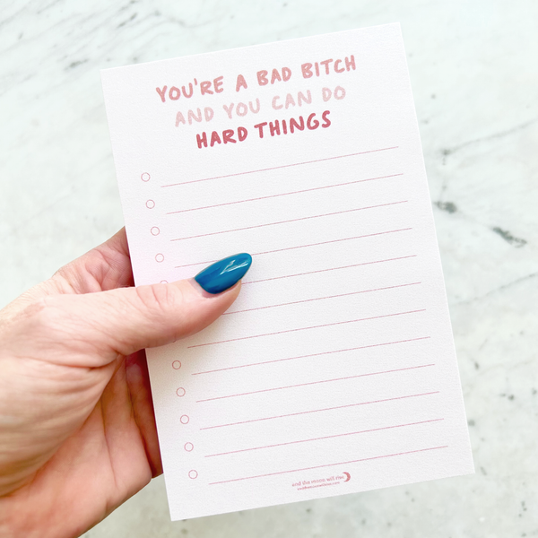 hand holding 4"W x 6"L sticky notepad; "you're a bad bitch and you can do hard things" in pink font at top with 12 horizontal light pink lines for list making on light pink background