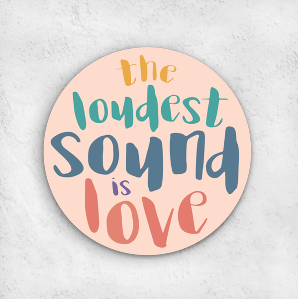 3" round waterproof vinyl sticker "the loudest sound is love" in gold, teal, blue, purple and coral funky font on a pink background shown on gray surface