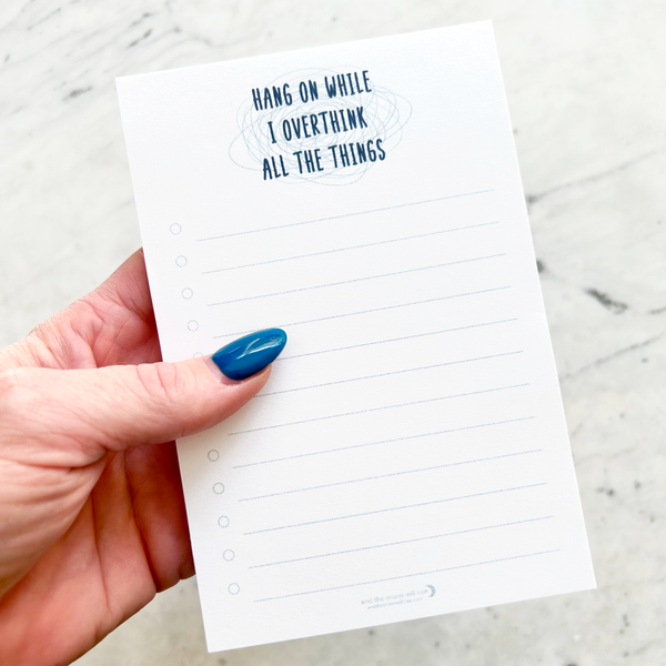 hand holding 4"W x 6"L sticky notepad; "hang on while I overthink all the things" in navy font atop light blue chaos squiggles at top with 12 horizontal light blue lines for list making on white background