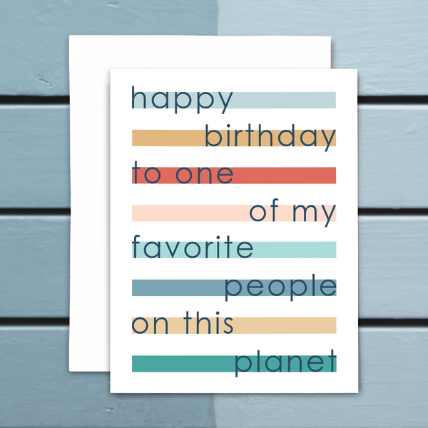 Greeting card "happy birthday to one of my favorite people on this planet"; colorful horizontal stripes under navy blue font on white background; blank interior with white envelope; shown on blue wood background