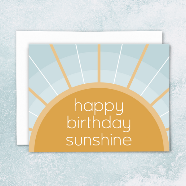 blank greeting card, "happy birthday sunshine" in white sans-serif font on golden sun with rays over concentric blue arches with white envelope on blue watercolor background