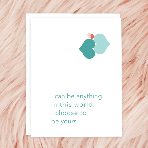 "I can be anything in this world. I choose to be yours." greeting card; pink and teal hearts with teal sans serif font on white background; blank interior with white envelope. Shown photographed on light pink faux fur.