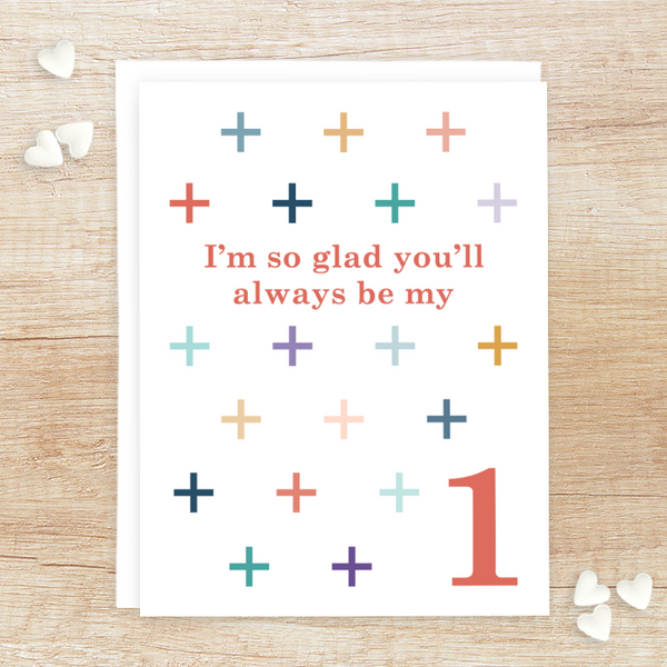 "I'm so glad you'll always be my +1" greeting card; coral serif font surrounded by grid of + in various colors (blues, teals, golds, corals, pinks, purples) on white background; blank interior with white envelope. Shown photographed on wood background with white candy hearts.