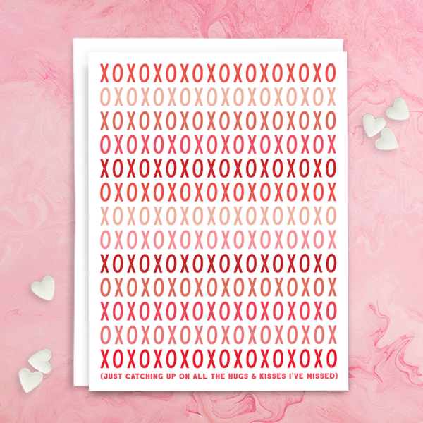 "XOXOXOXOXO Just catching up on all the hugs & kisses I've missed" greeting card; font in shades of reds and pinks on white background; blank interior with white envelope. Shown photographed on pink background with white candy hearts.