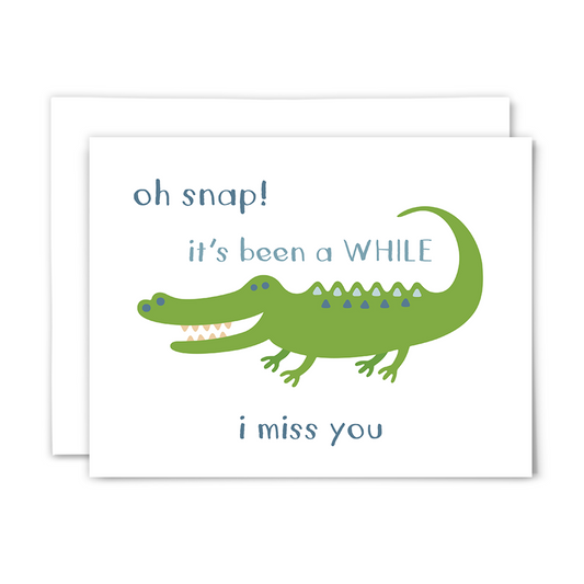 Folded A2 greeting card with blank interior and white envelope; front reads ""oh snap! it's been a WHILE ~ i miss you" with a big, green, cartoon crocodile with a big toothy smile on a white background