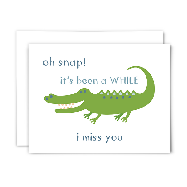 Folded A2 greeting card with blank interior and white envelope; front reads ""oh snap! it's been a WHILE ~ i miss you" with a big, green, cartoon crocodile with a big toothy smile on a white background