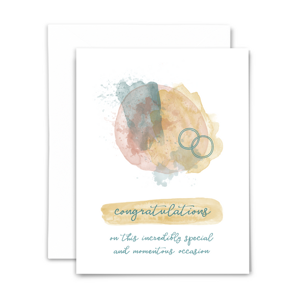 A white greeting card with wedding bands and text 'congratulations on this incredibly special and momentous occasion' in script font. The card is accompanied by a white envelope.