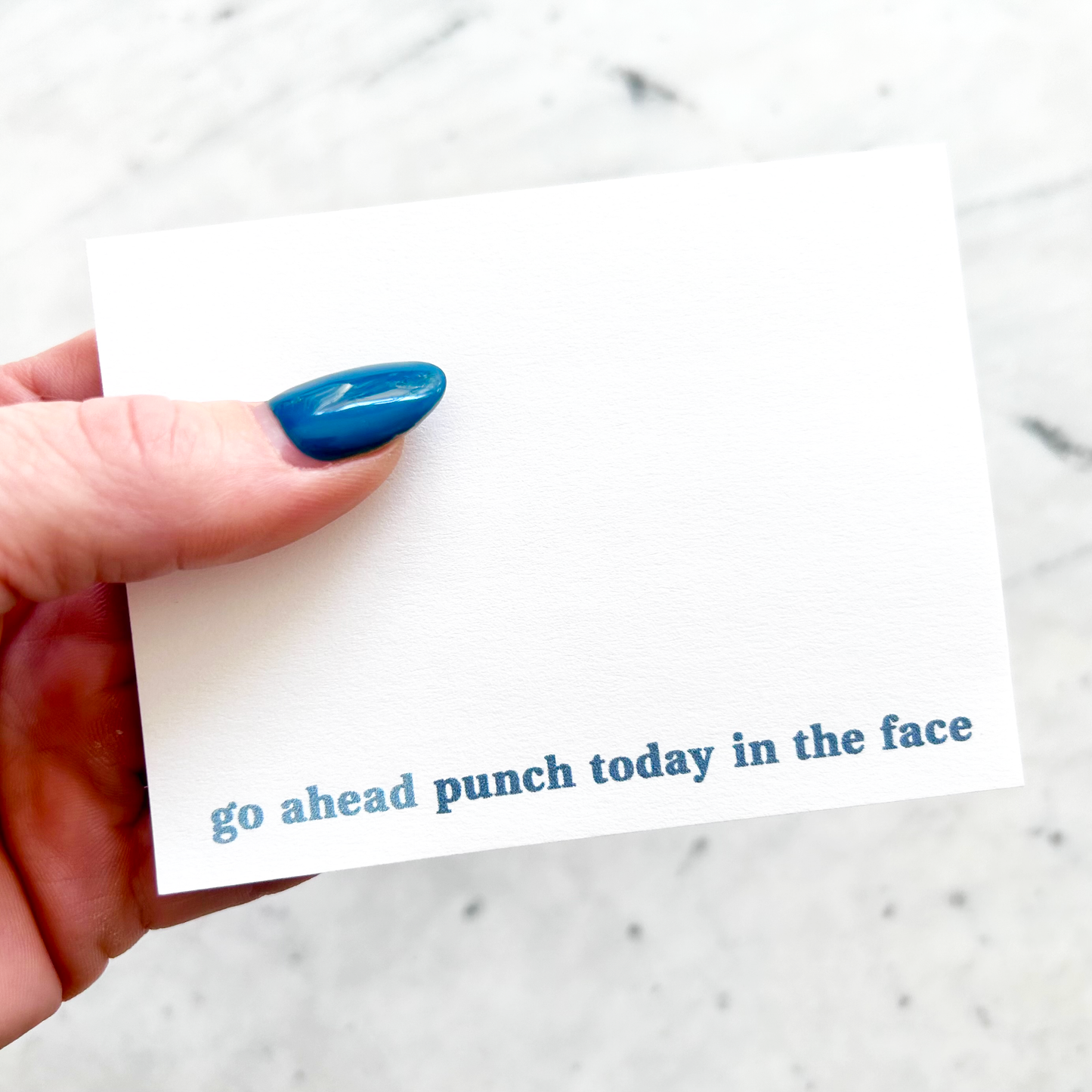 hand holding 4"W x 3"L sticky notepad; "go ahead ~ punch today in the face" in blue lowercase font along bottom on white background