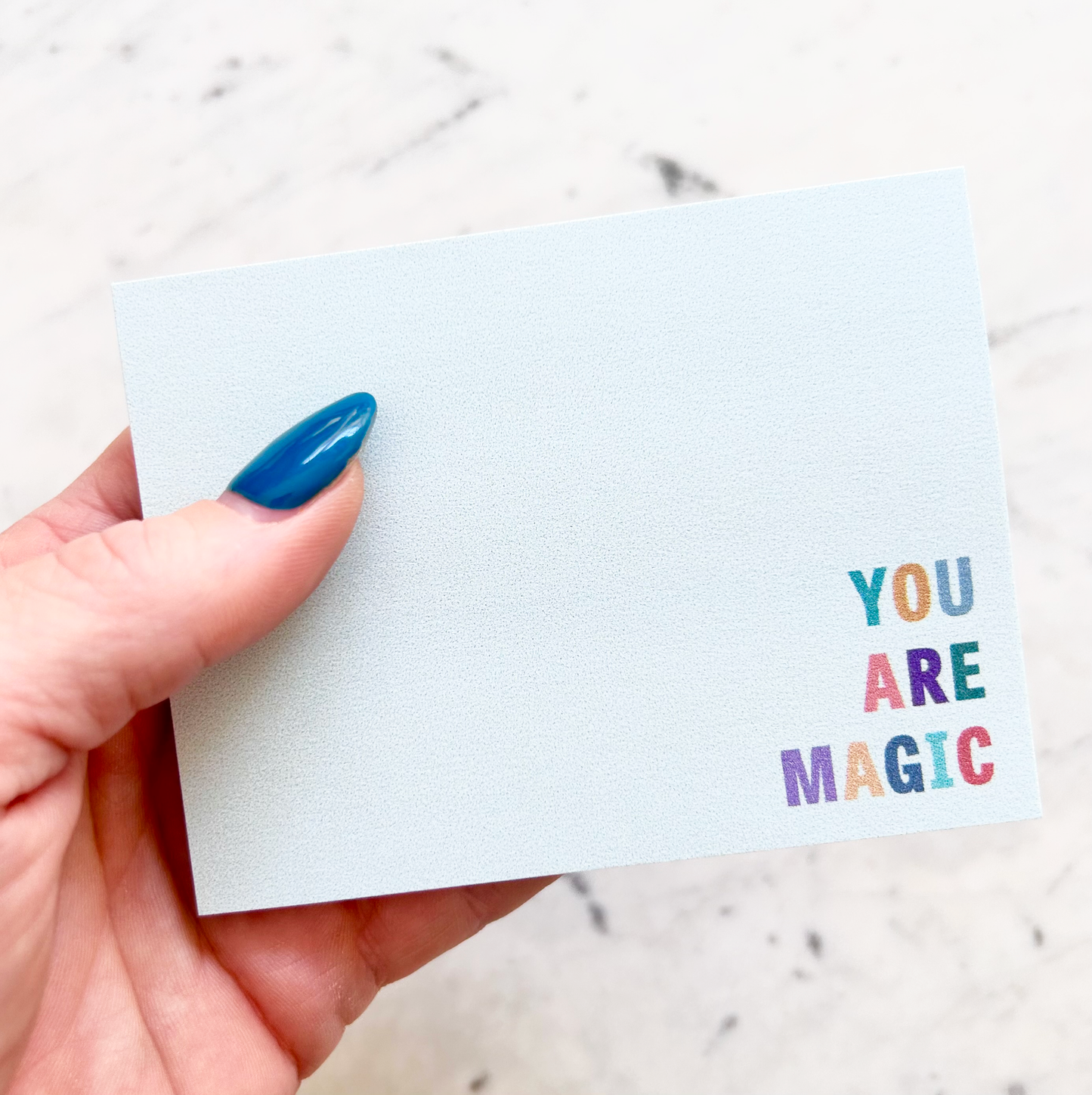 hand holding 4"W x 3"L sticky notepad; "you are magic" in colorful block letters in lower right corner on light teal background