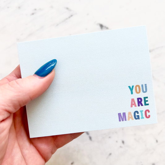 hand holding 4"W x 3"L sticky notepad; "you are magic" in colorful block letters in lower right corner on light teal background