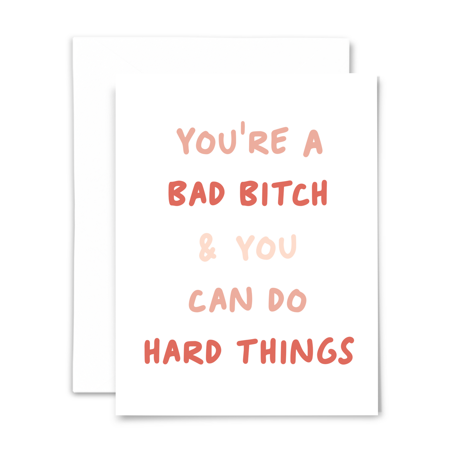 A white greeting card with pink and coral handwritten font 'You're a bad bitch and you can do hard things'. The card is accompanied by a white envelope.