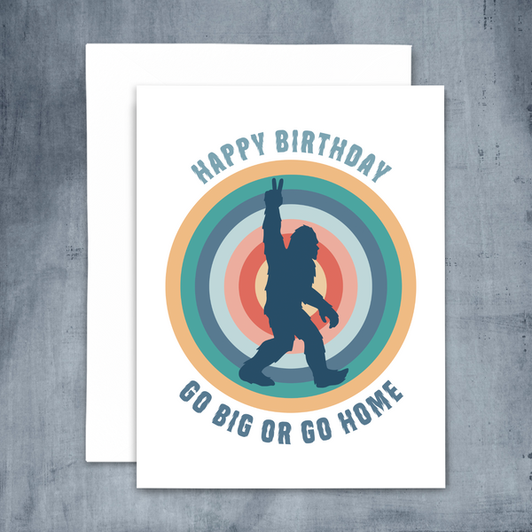 blank greeting card "happy birthday go big or go home" in hairy blue font with navy bigfoot silhouette over colorful concentric circles on blue concrete background with white envelope