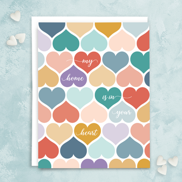 "My home is in your heart" greeting card; white script font on top of patterned hearts in various colors- blues, teals, golds, pinks, purples; white interior with white envelope. Shown photographed on light blue background with white candy hearts.